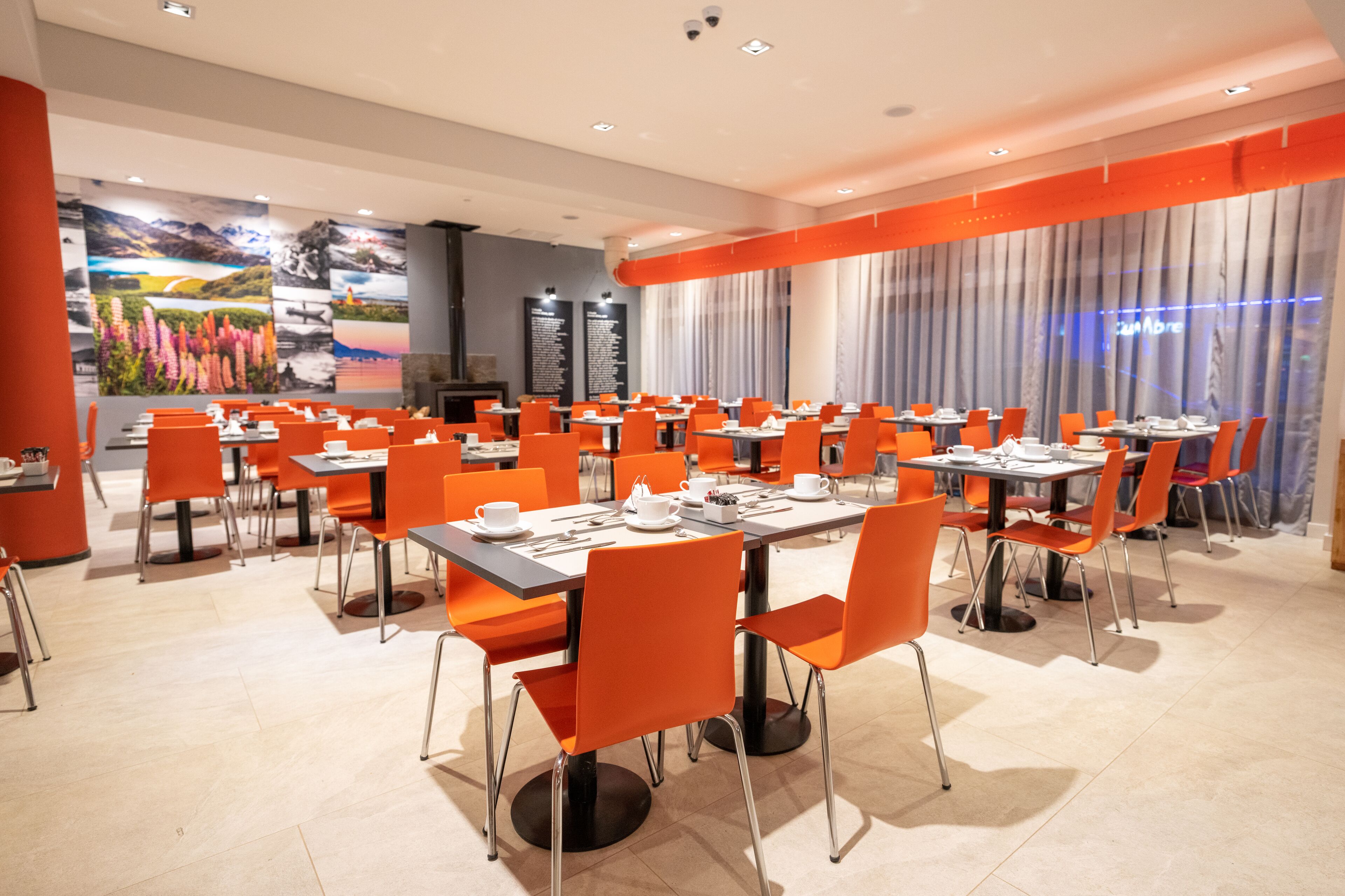 A dining area with multiple tables and orange chairs, featuring a large mural on the wall and a view of the cityscape through large windows.