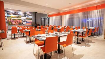 A dining area with multiple tables and orange chairs, featuring a large mural on the wall and a view of the cityscape through large windows.