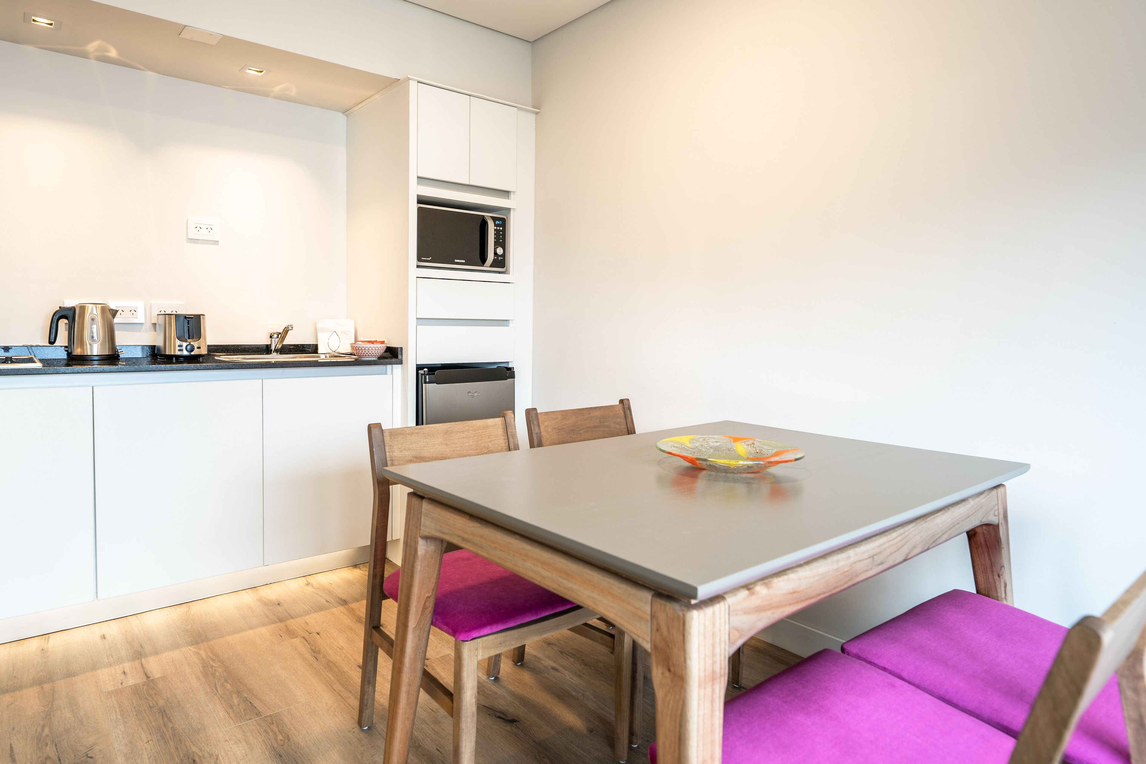 A compact kitchen with white cabinets, a microwave, and a kettle. There is a dining table with two chairs, one with a purple cushion.