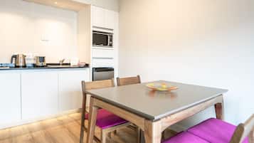 Una cocina compacta con armarios blancos, un microondas y una tetera. Hay una mesa de comedor con dos sillas, una con cojín morado.