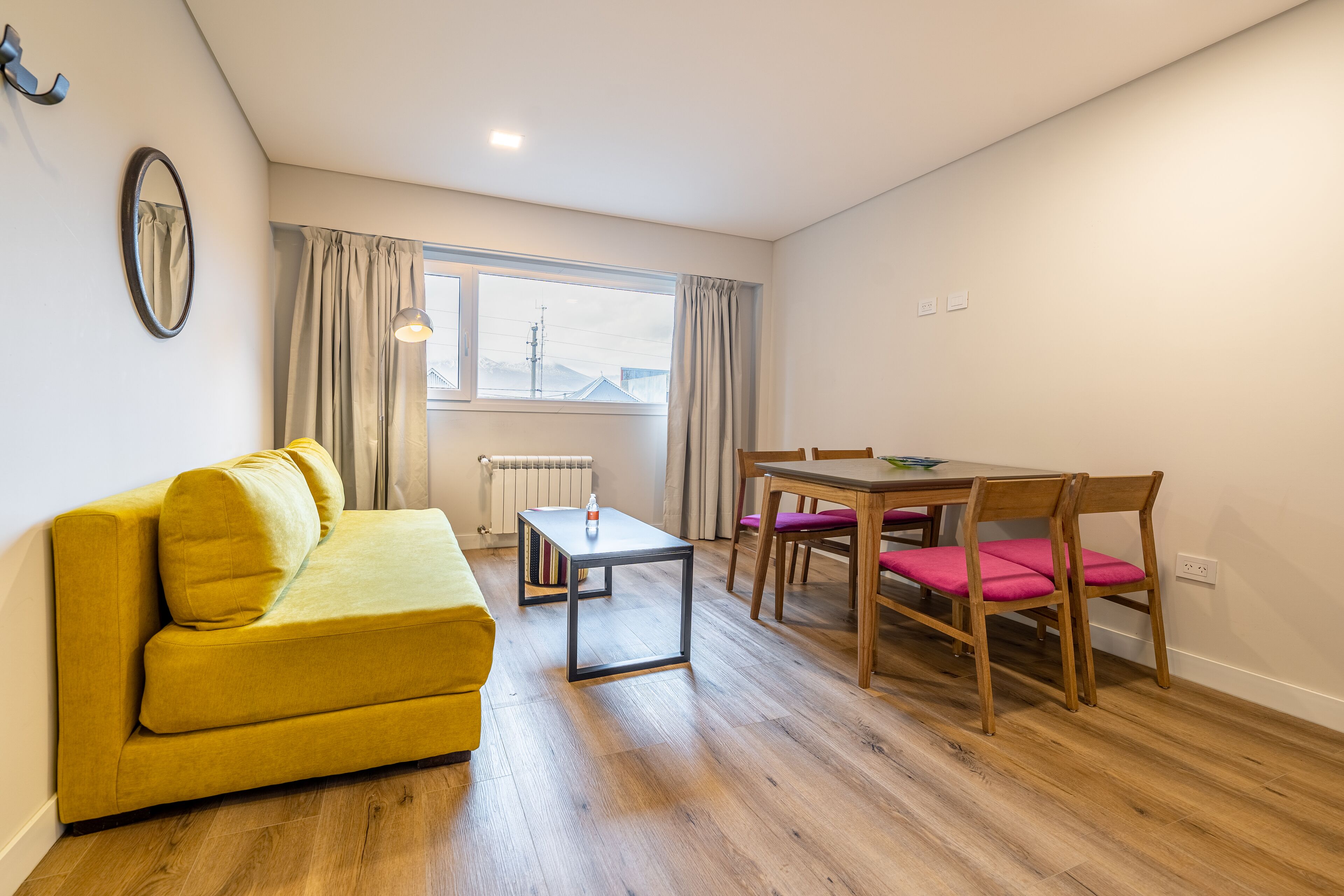 A compact living space with a yellow sofa, a wooden dining table with chairs, a small side table, and a window with curtains.