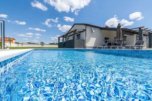 Una piscina con acqua cristallina, un'area patio coperta con poltrona relax e un edificio con facciata bianca sullo sfondo.