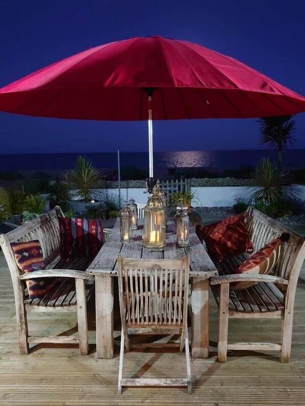 Une terrasse en bois avec un parasol rouge, une table et des chaises. On aperçoit des lanternes sur la table et, au loin, une vue sur l’océan.