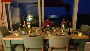 Een eettafel gedekt voor vier personen, met kaarsen, glazen en flessen, met uitzicht op een schilderachtig zeegezicht tijdens de zonsondergang.