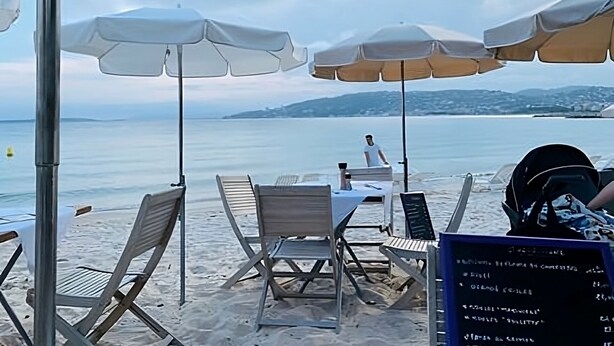 Un café frente a la playa con terraza, incluyendo sillas y sombrillas, y un cartel con la carta visible.