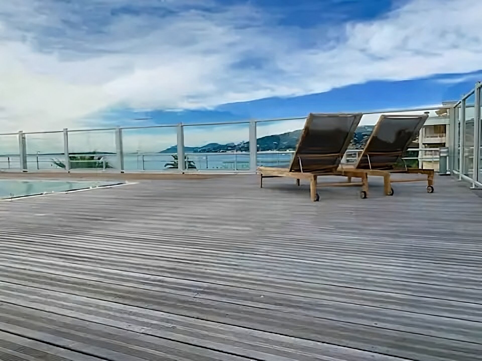 A wooden deck with two sun loungers and a glass railing overlooking a body of water and distant mountains.