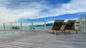 Terraza de madera con dos tumbonas y barandilla de cristal con vista a un cuerpo de agua y montañas distantes.