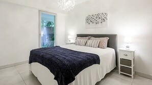 Un dormitorio moderno con una cama grande, mesitas de noche, una araña y vistas al verdor por la ventana.