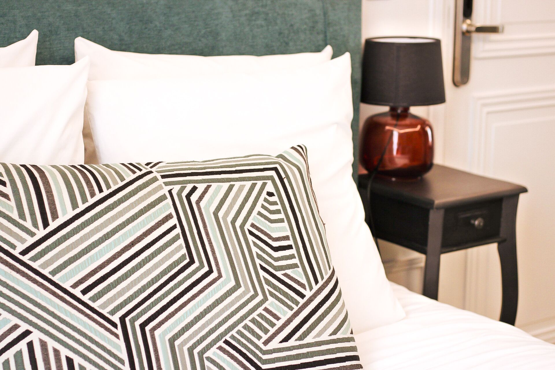 A neatly made bed with white bedding and a patterned throw pillow. A small bedside table with a lamp and a red vase is visible.