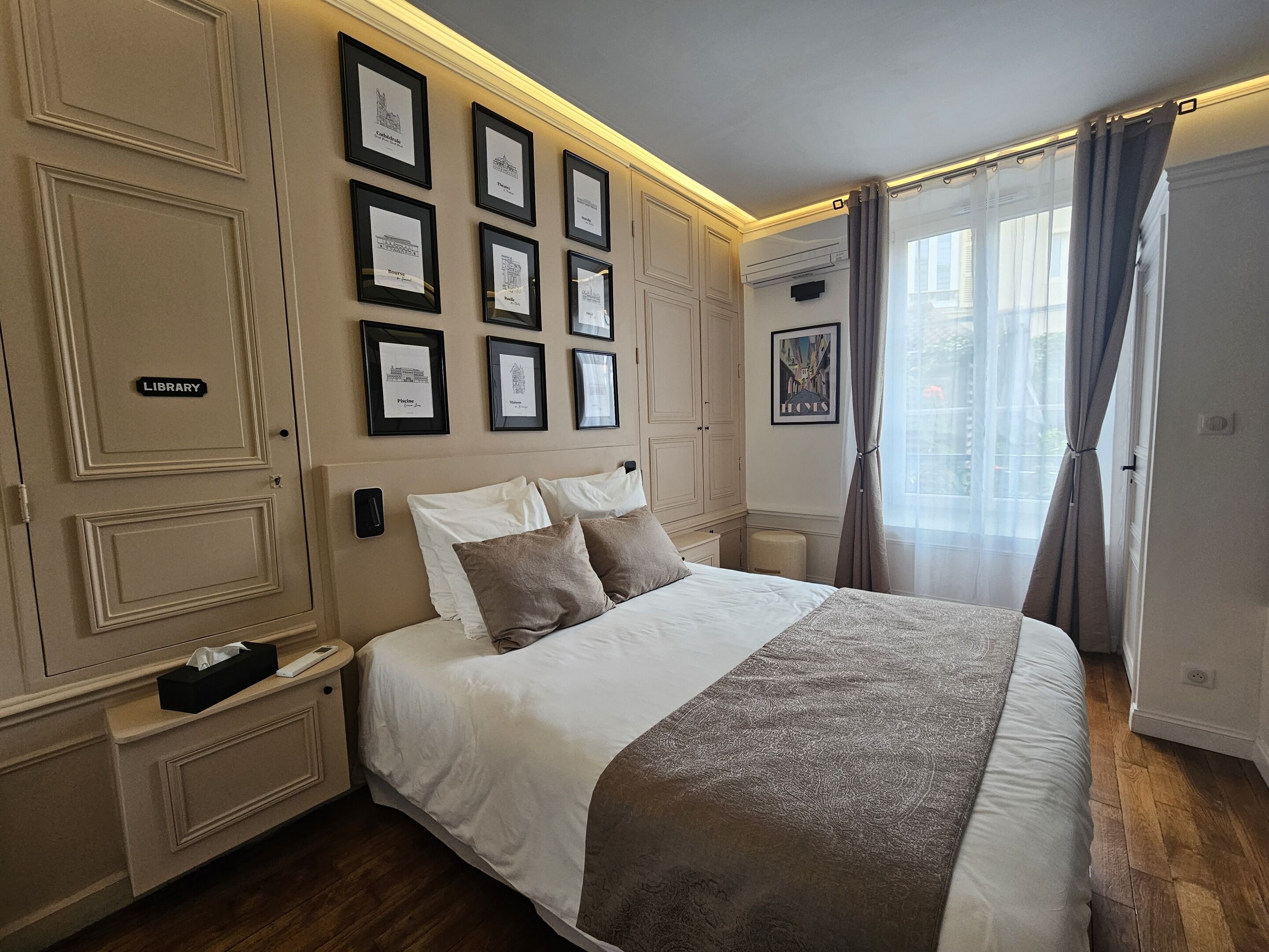 A hotel room with a large bed, a nightstand, a bedside table, a wardrobe, and a wall with framed pictures.