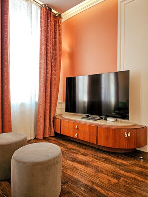 Ein modernes Wohnzimmer mit einem Flachbildfernseher auf einer Holzkommode, zwei Hocker und einem großen Fenster mit roten Vorhängen.