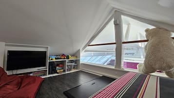 A room with a sloped ceiling, a large window, a television, a sofa, and a teddy bear.