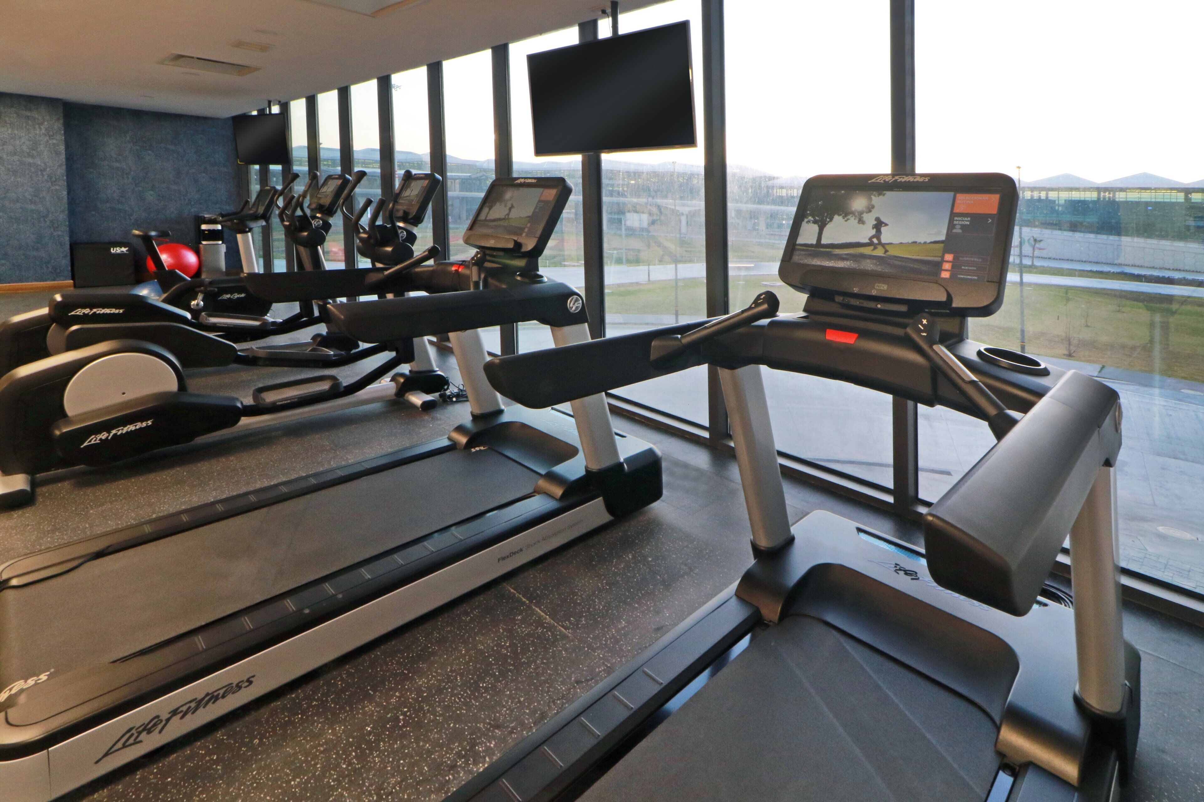 A gym with treadmills, stationary bikes, and a rowing machine, with large windows offering a view of a bridge and landscape.