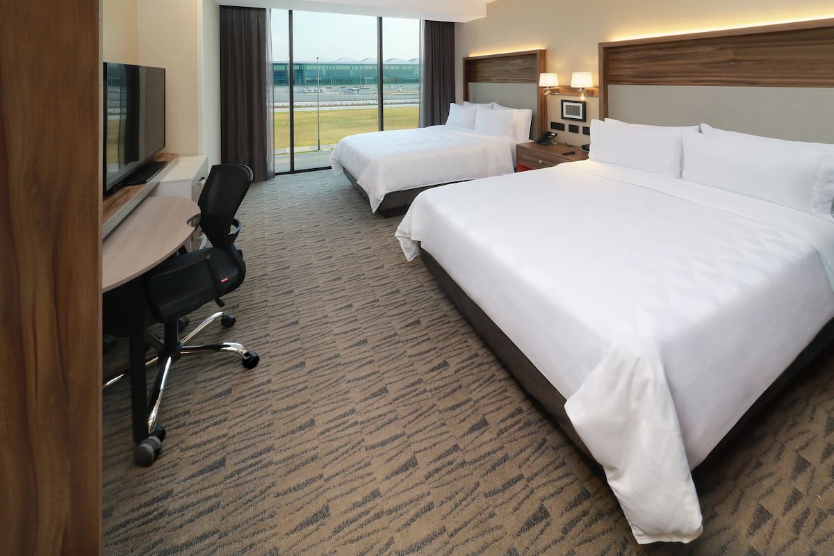 Une chambre d’hôtel avec deux lits, un bureau avec une télévision, une chaise et une vue sur un aéroport.