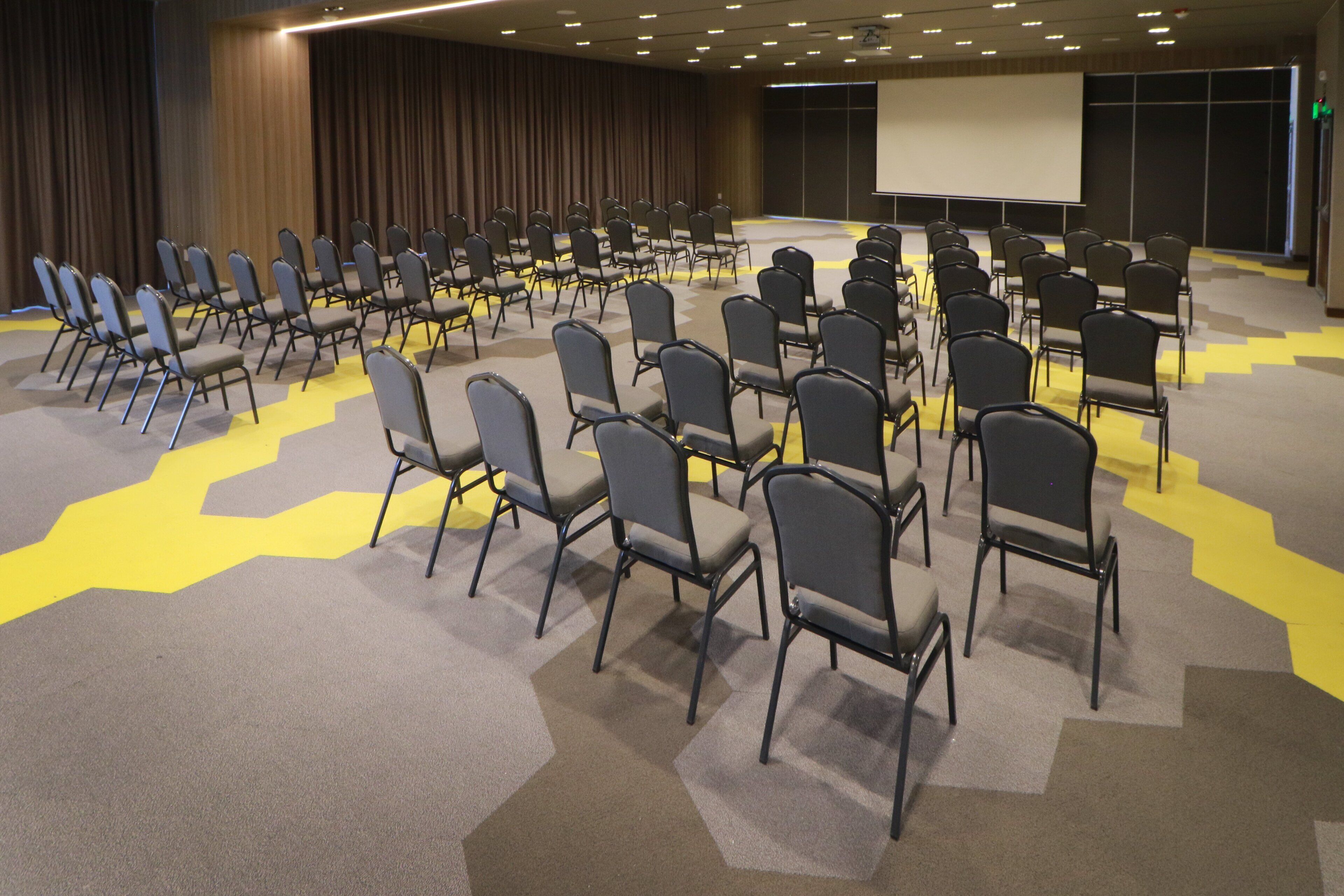 Un salón de conferencias con filas de sillas orientadas hacia una pantalla de proyección, suelo alfombrado con diseños geométricos y una zona de escenario.