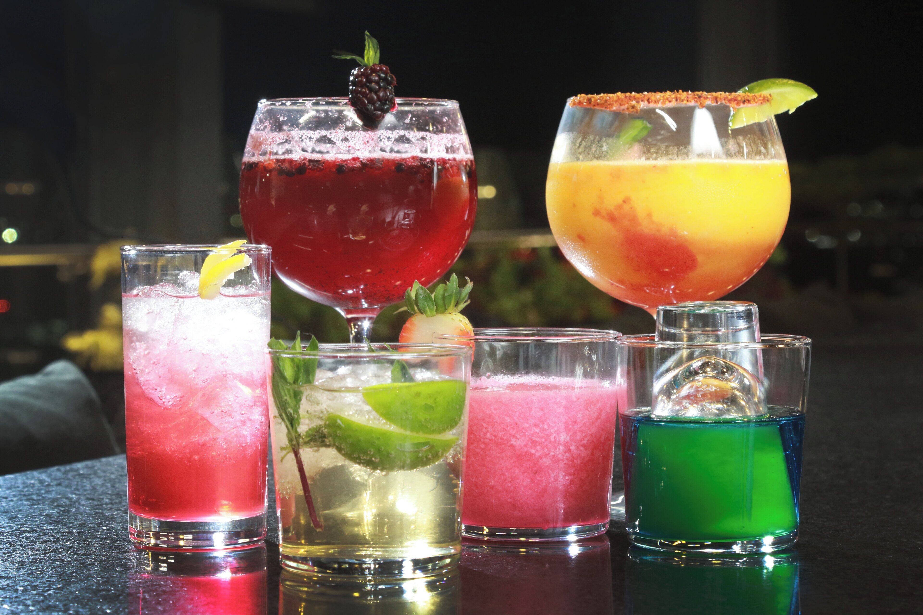 Variedad de cócteles coloridos en distintos vasos, algunos adornados, sobre una barra.