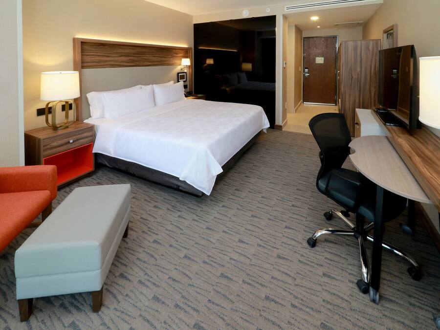 Habitación de hotel con una cama grande, un escritorio con silla, un sillón naranja pequeño y un televisor.