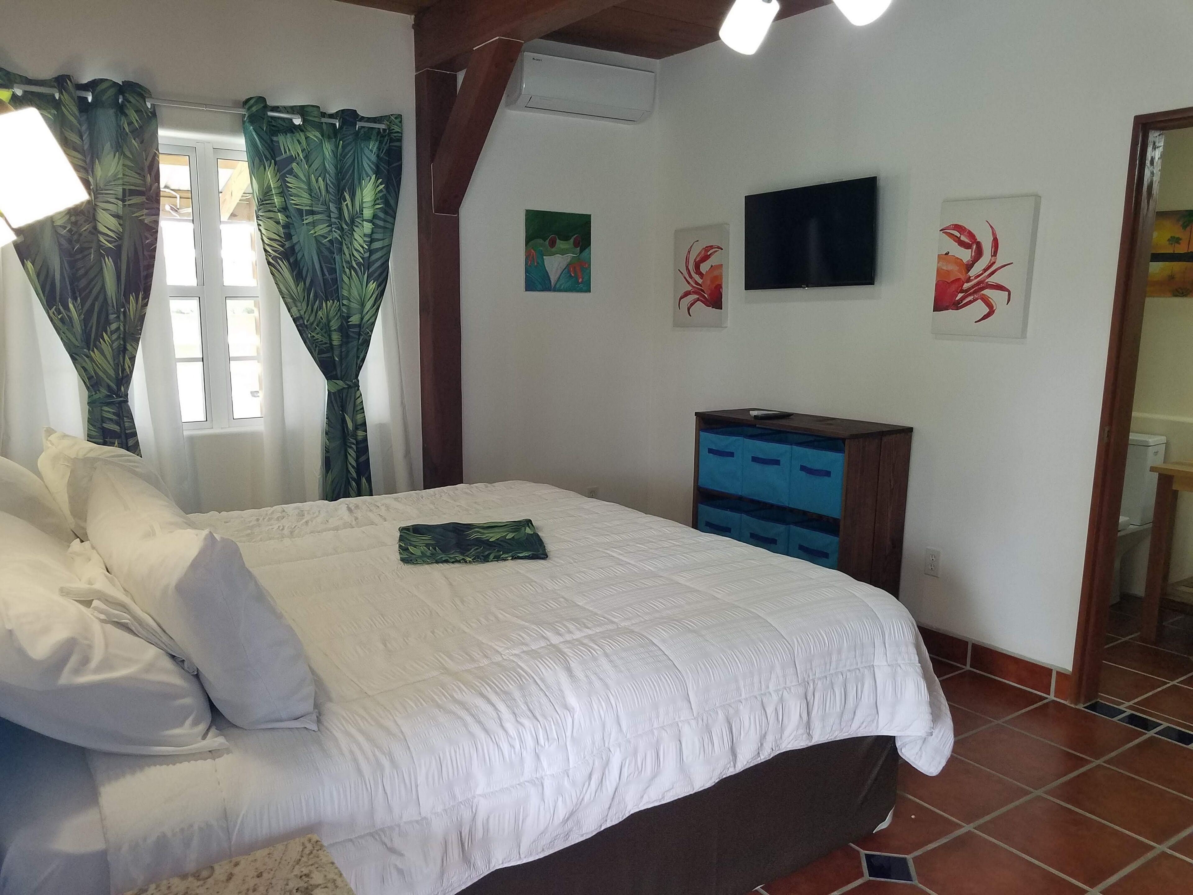 Une chambre à coucher comprenant un lit, des rideaux, un téléviseur, des œuvres d’art et un meuble de rangement.