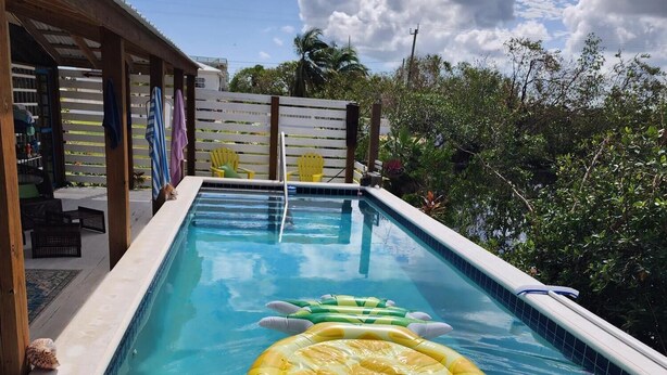 Una piscina con agua azul cristalina, un flotador amarillo y una zona de madera con sillas y una valla.