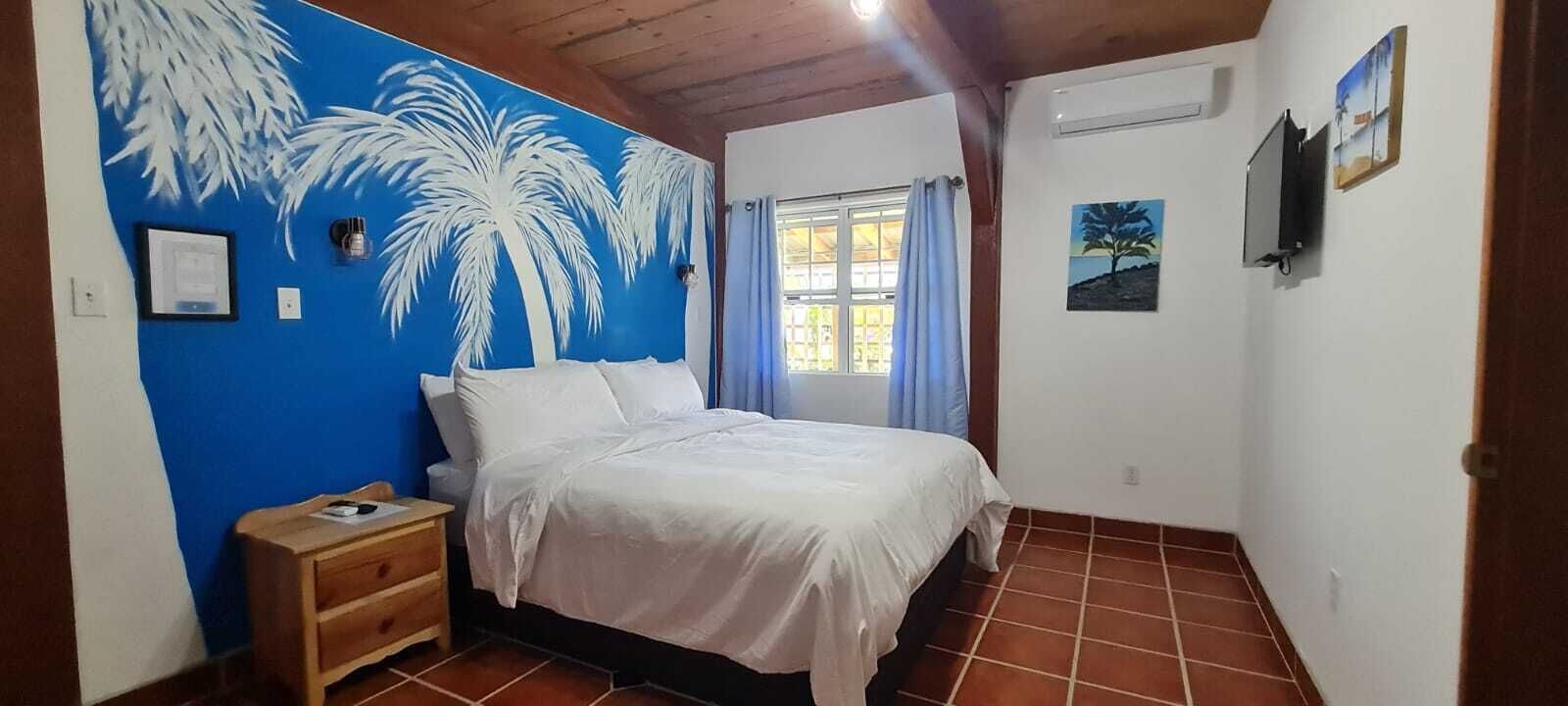 A bedroom with a bed, a nightstand, a window with curtains, and a wall with palm tree artwork.