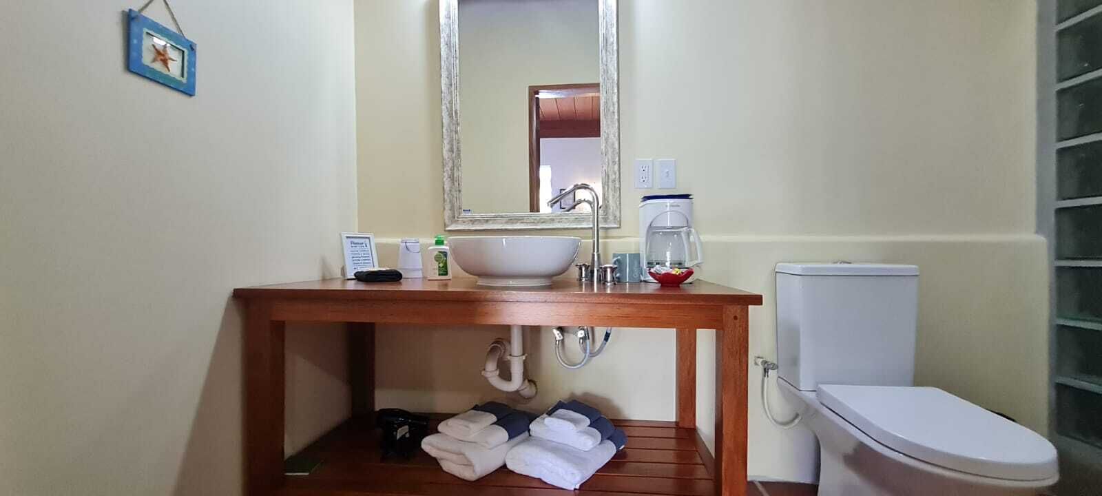 Un baño con tocador de madera, lavamanos blanco, espejo, inodoro y repisa con toallas.