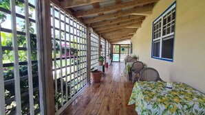 Un corredor cubierto con suelo de madera, una mesa con mantel floral, plantas en macetas y una ventana con marco azul.