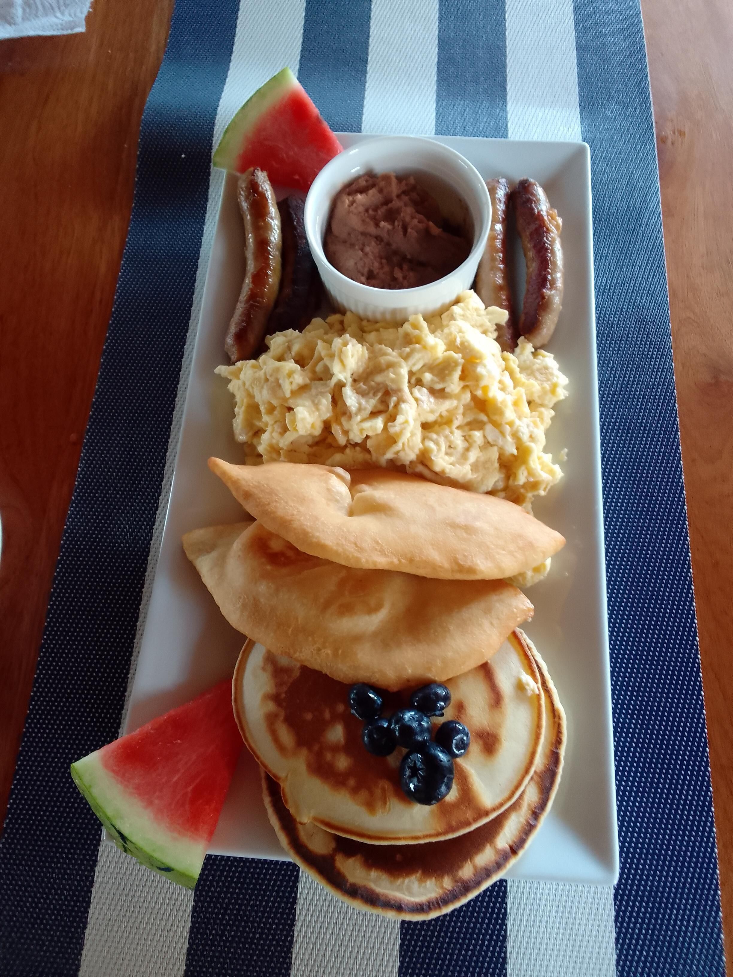 A breakfast plate with pancakes, sausage, scrambled eggs, and a small bowl of sauce.