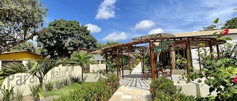 Un sendero en el jardín que conduce a una pérgola de madera con asientos, rodeada de abundante vegetación y plantas en flor.