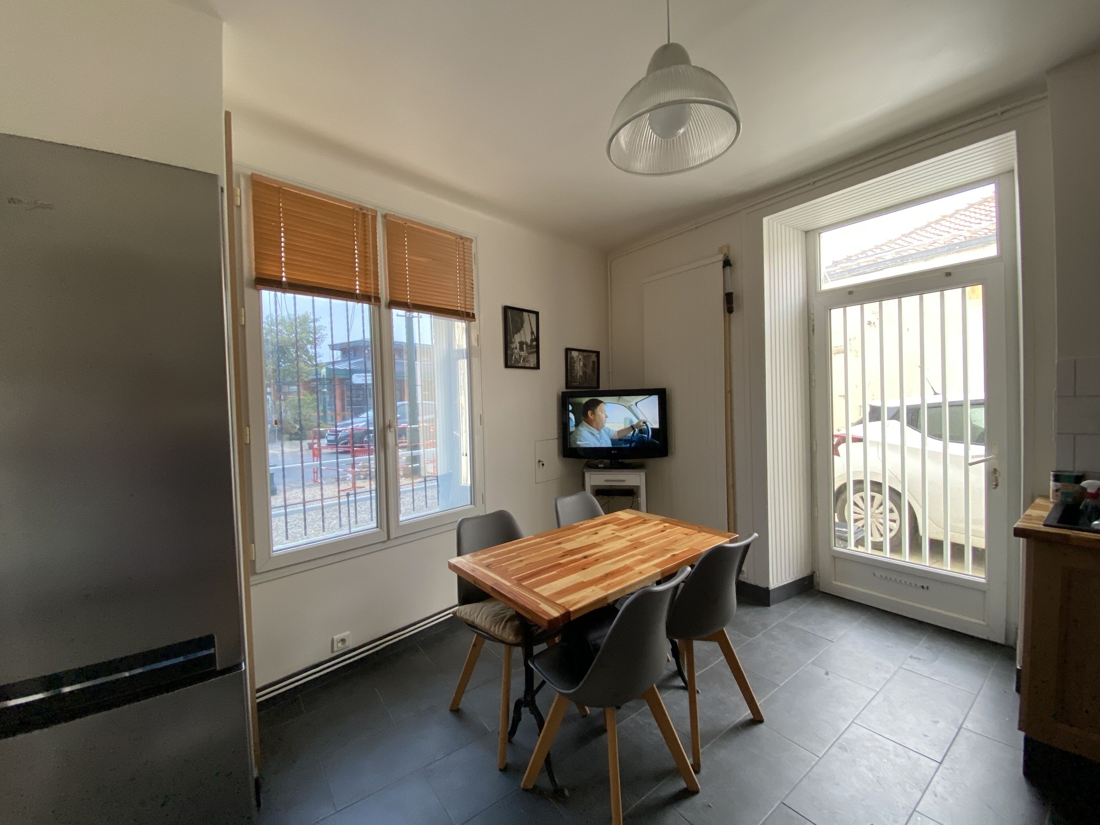 A dining area with a wooden table and chairs, a television on a stand, and large windows with blinds.