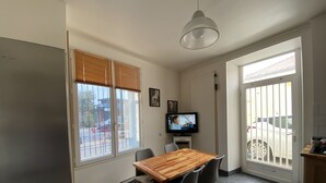 A dining area with a wooden table and chairs, a television on a stand, and large windows with blinds. - SEASONAL HOUSE RENTAL (Les Sables-d'Olonne)