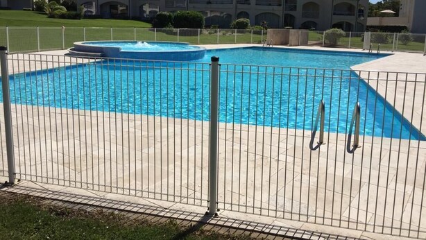 Ein rechteckiger Swimmingpool mit einer Fontäne, umgeben von einem Metallzaun. Im Hintergrund sind mehrstöckige Gebäude mit Balkonen zu sehen.