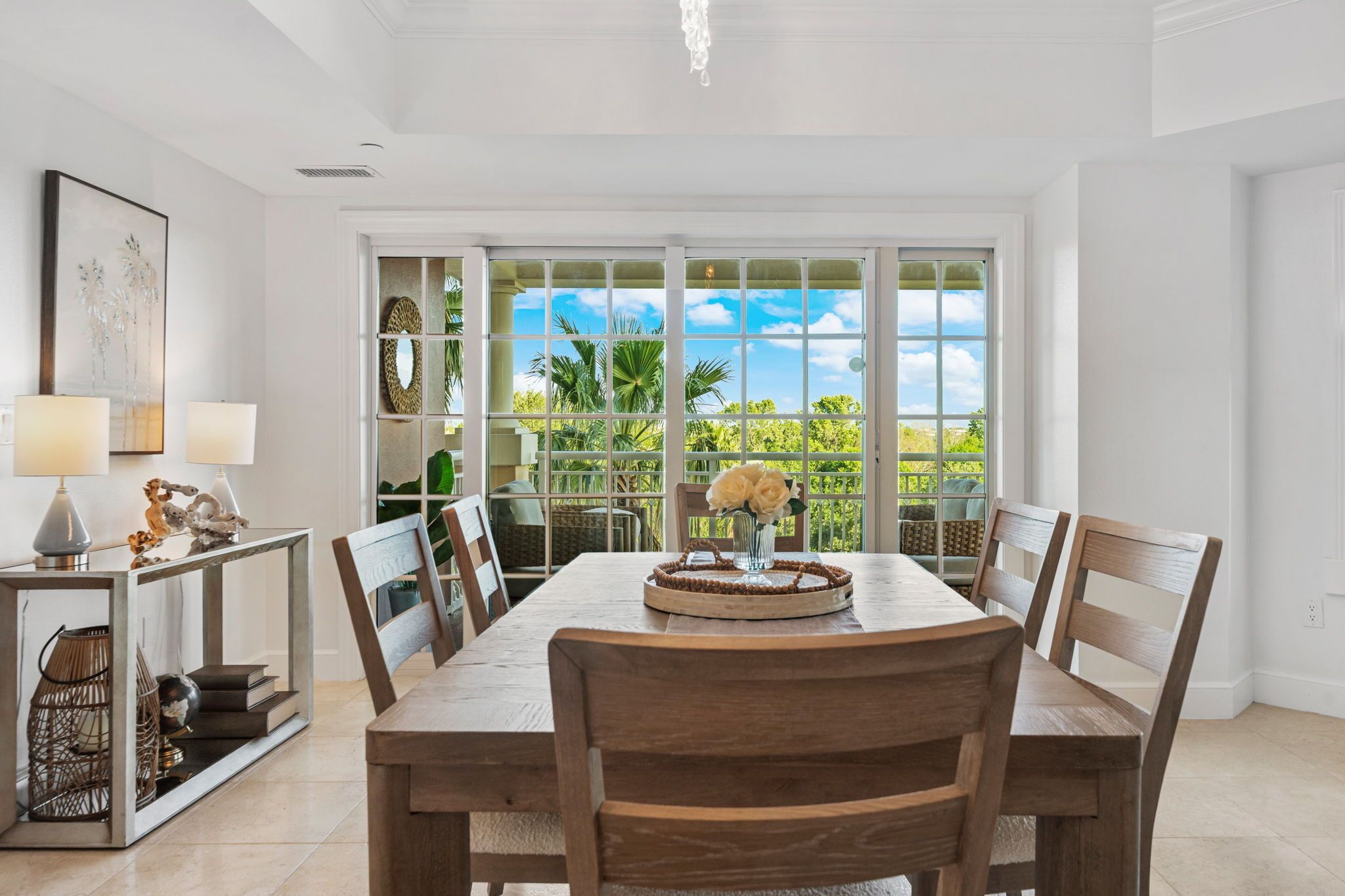 A dining area with a large table, chairs, a vase of flowers, and a view through a glass door to the outdoors.