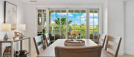 A dining area with a large table, chairs, a vase of flowers, and a view through a glass door to the outdoors.