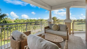 Ein überdachter Balkon mit Korbmöbeln, einer gepolsterten Couch und Blick auf einen Golfplatz.