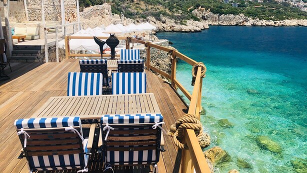 A wooden deck with striped lounge chairs and a table, overlooking a clear blue sea with rocky terrain and buildings in the distance. - La Mer Comfort Kalkan (Kalkan - Kaş)