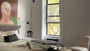 Een slaapkamer met een groot raam, een bed met dekbed, een stoel en een schilderij aan de muur.