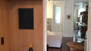 Een gang met een houten plank waarop boeken, een fles wijn en glazen staan. Achtergrond toont een witte bank en een keuken.