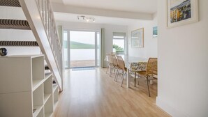 A spacious room with a wooden floor, a dining area with wicker chairs, a staircase, and a view of the outdoors. - 5 Bed in Woolacombe (oc-bella) (Woolacombe)