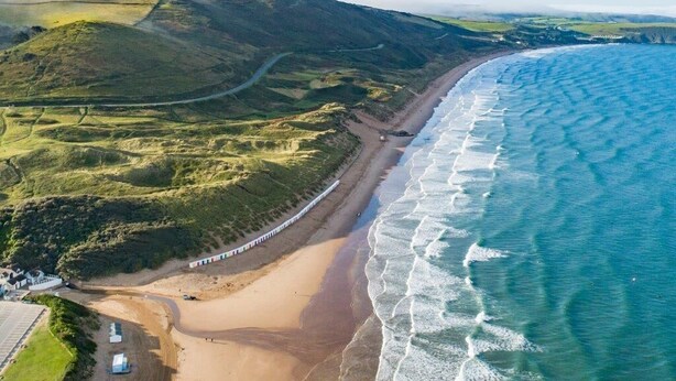 An aerial view of a coastal area with a beach, rolling hills, and a road curving along the coastline. - 5 Bed in Woolacombe (oc-bella) (Woolacombe)