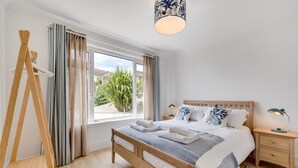 A bedroom with a wooden bed, white bedding, a bedside table with lamps, a window with curtains, and a view of greenery outside. - 5 Bed in Woolacombe (oc-bella) (Woolacombe)