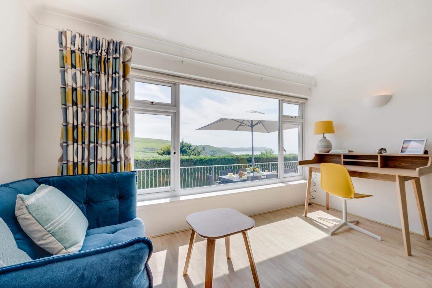 Quarto moderno com sofá azul, uma pequena mesa branca, uma cadeira amarela, escrivaninha de madeira e uma janela grande com vista para o verde.