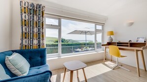 A modern room with a blue sofa, a small white table, a yellow chair, a wooden desk, and a large window with a view of greenery. - 5 Bed in Woolacombe (oc-bella) (Woolacombe)