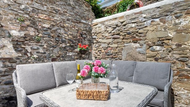 A wicker table with a grey cushion, a basket, wine glasses, a bottle, and a vase with flowers.