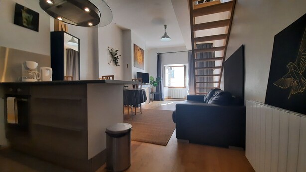 A modern living room with a dark sofa, a coffee table, and a TV. There's a kitchen area with a counter and a sink. A ladder is visible against the wall, and a window lets in natural light.