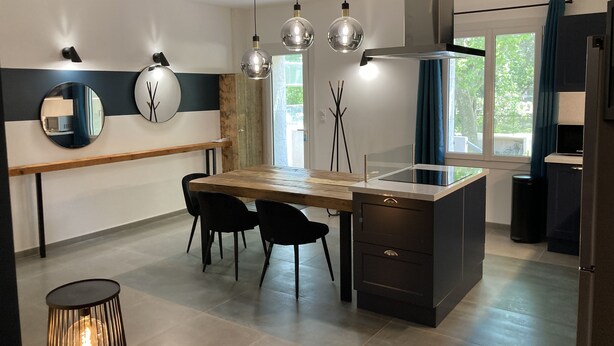 Une cuisine moderne avec une table en bois, des chaises noires, un îlot central intégré et des luminaires suspendus.
