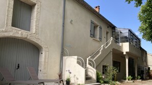 Un bâtiment à la façade beige, avec un escalier menant à un balcon et un petit jardin agrémenté d’un banc et de plantes en pot.