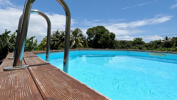 Une terrasse en bois avec un escalier métallique menant à une piscine à l’eau d’un bleu limpide, le tout entouré d’une végétation luxuriante.