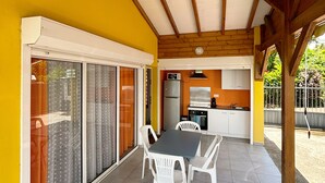 Une terrasse couverte avec une table et des chaises, un coin cuisine et une porte-fenêtre coulissante donnant sur un espace extérieur.