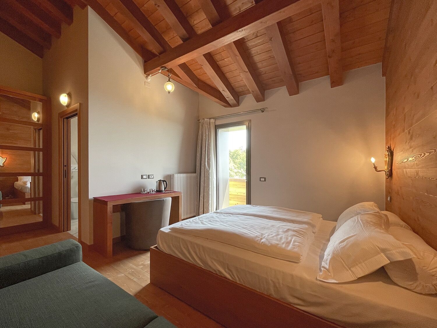 A bedroom with a wooden ceiling, a bed with white linens, a desk with a kettle, and a view through a window.
