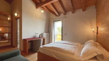 A bedroom with a wooden ceiling, a bed with white linens, a desk with a kettle, and a view through a window.